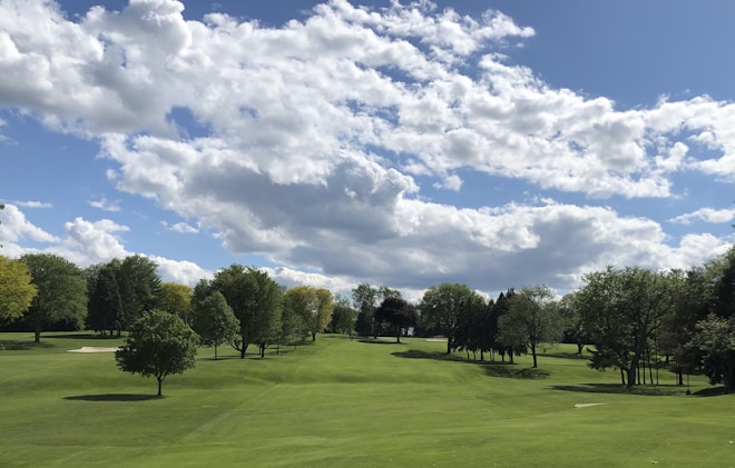 A lush green golf course with neatly trimmed grass, surrounded by a variety of trees under a bright blue sky filled with scattered white clouds.