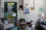 A cozy living room with stylish decor, featuring two armchairs, a sofa, various artworks on the walls, and a collection of potted plants. There are two lamps flanking the seating area, with one larger artwork displaying a waterfall scene. Shelves hold books and audio speakers, and a decorative antler piece is mounted above the central wall art.