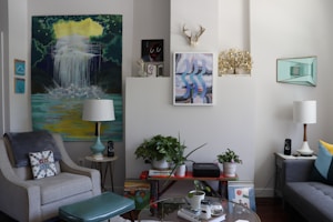 A cozy living room with stylish decor, featuring two armchairs, a sofa, various artworks on the walls, and a collection of potted plants. There are two lamps flanking the seating area, with one larger artwork displaying a waterfall scene. Shelves hold books and audio speakers, and a decorative antler piece is mounted above the central wall art.
