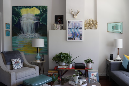 A cozy living room with stylish decor, featuring two armchairs, a sofa, various artworks on the walls, and a collection of potted plants. There are two lamps flanking the seating area, with one larger artwork displaying a waterfall scene. Shelves hold books and audio speakers, and a decorative antler piece is mounted above the central wall art.