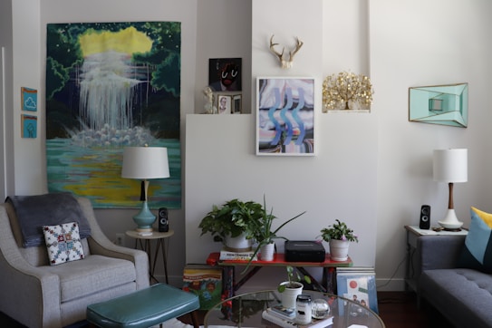 A cozy living room with stylish decor, featuring two armchairs, a sofa, various artworks on the walls, and a collection of potted plants. There are two lamps flanking the seating area, with one larger artwork displaying a waterfall scene. Shelves hold books and audio speakers, and a decorative antler piece is mounted above the central wall art.