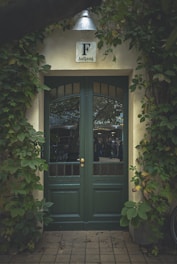 A green wooden door with glass panels is framed by ivy-covered walls. Above the door, there is a metal lamp and a sign with the letter 'F' and the word 'Aufgang'. The scene appears to be at the entrance of a building, possibly in a garden or courtyard setting.