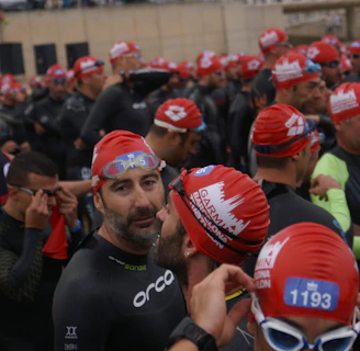 A large group of people wearing swim caps and wetsuits, appearing to be participants in a swimming event. They are gathered closely together, some adjusting their gear, with a variety of expressions on their faces.