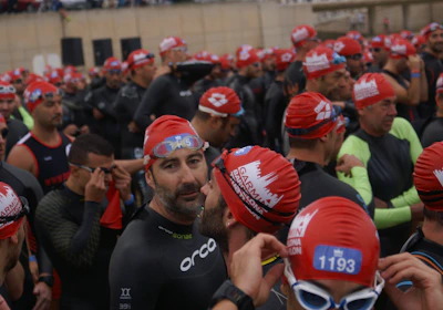 A large group of people wearing swim caps and wetsuits, appearing to be participants in a swimming event. They are gathered closely together, some adjusting their gear, with a variety of expressions on their faces.