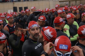 A large group of people wearing swim caps and wetsuits, appearing to be participants in a swimming event. They are gathered closely together, some adjusting their gear, with a variety of expressions on their faces.