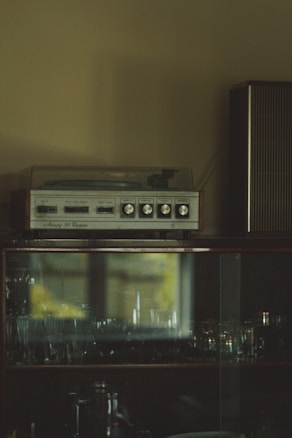 A vintage stereo system sits atop a dark wooden cabinet with glass doors, reflecting light and glassware inside. The room has a dimly lit atmosphere, with shadows and muted tones.