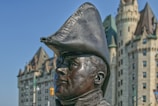 A bronze statue of a historical figure wearing a distinctive tricorn hat stands prominently. Behind the statue, an ornate stone building with turrets and towers is visible, suggesting a historical or architectural significance.