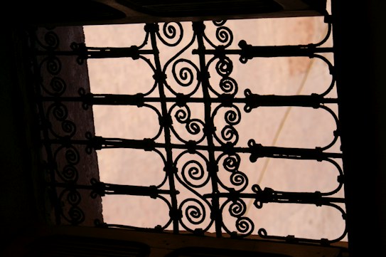 An intricately designed wrought iron gate pattern with swirling motifs is silhouetted against a soft, pastel-colored background. The ornamental design features symmetrical and geometric shapes, giving it a classic and artistic appearance.