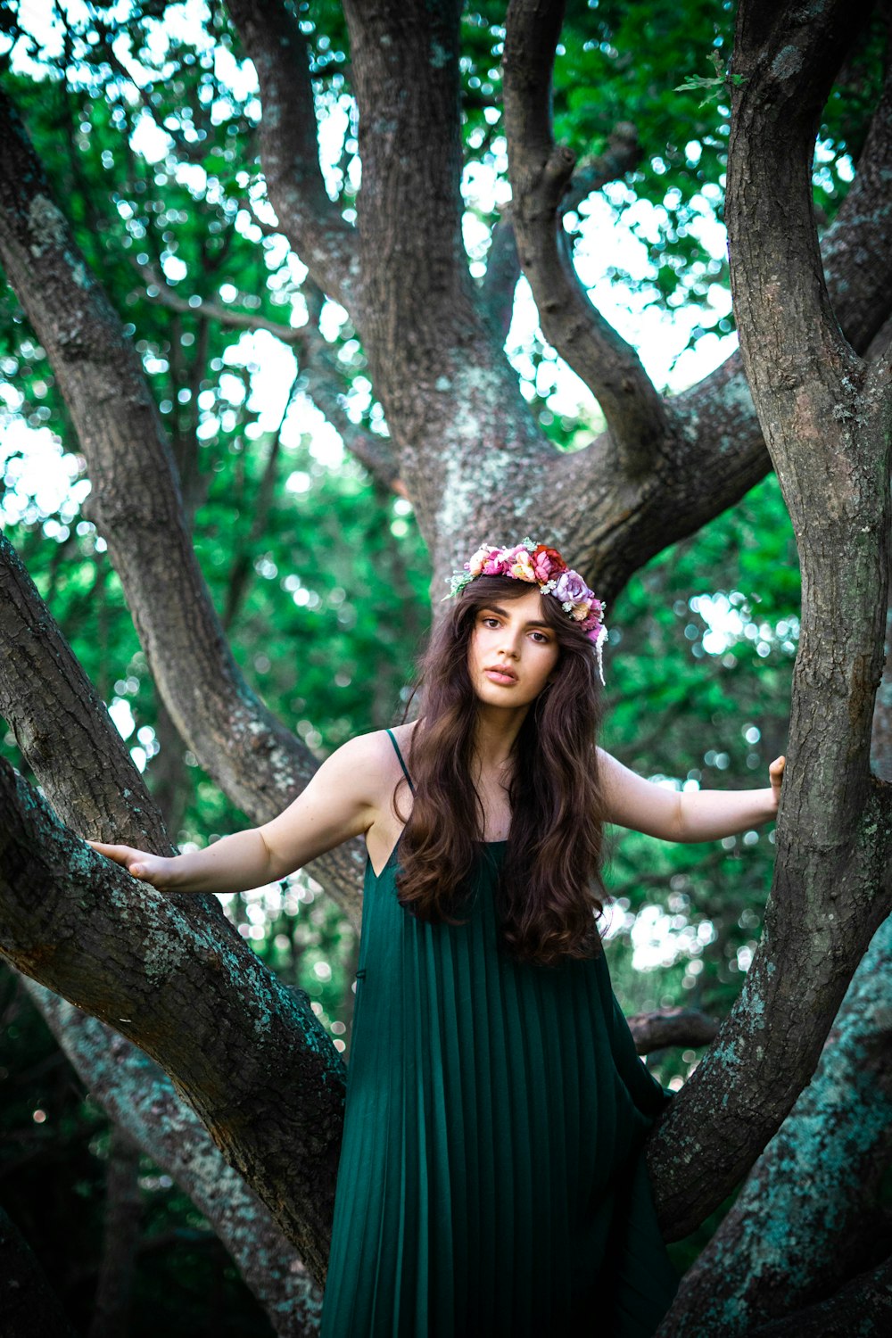 woman in green sleeveless dress wearing pink floral headdress standing beside brown tree