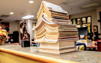 A tall stack of books is neatly arranged on a counter in a warmly lit room. The books vary in thickness and cover color, creating a visually interesting pattern. The background features blurred office elements, such as computers and framed items on the walls, suggesting an educational or work environment.