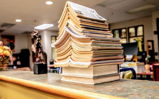 A tall stack of books is neatly arranged on a counter in a warmly lit room. The books vary in thickness and cover color, creating a visually interesting pattern. The background features blurred office elements, such as computers and framed items on the walls, suggesting an educational or work environment.