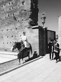 A person dressed in traditional attire is mounted on a horse, holding a lance. The horse has ornate decorations, and they are positioned beside a tall, ancient stone wall. Two other individuals are walking nearby, displaying casual attire. The scene is captured in black and white, enhancing the historic ambiance.