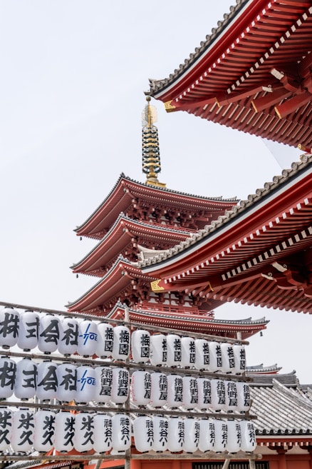 A traditional Japanese pagoda with intricate red and white architecture rises vertically, adorned with ornate details. Below, rows of white lanterns feature black Japanese characters, contributing to the cultural and historical ambiance.