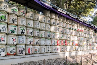 Rows of traditional Japanese sake barrels, called kazaridaru, are stacked on a wooden shelf under a canopy. Each barrel is decorated with colorful Japanese characters and symbols, representing various sake brewers and regions.