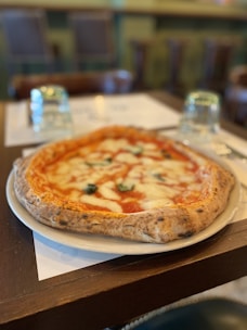 A delicious-looking pizza with a thick, golden-brown crust is placed on a white plate. The pizza is topped with melted mozzarella cheese, vibrant red tomato sauce, and fresh basil leaves. In the background, there are blurred images of chairs and a table, suggesting a cozy restaurant setting. Two upside-down glasses are placed on the table next to the plate.