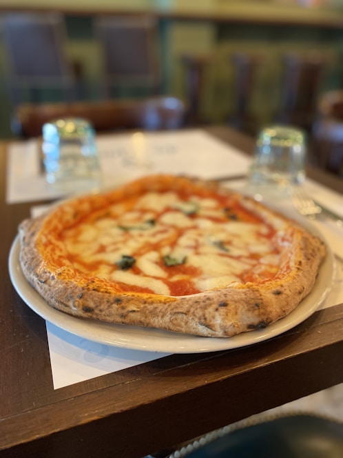 A delicious-looking pizza with a thick, golden-brown crust is placed on a white plate. The pizza is topped with melted mozzarella cheese, vibrant red tomato sauce, and fresh basil leaves. In the background, there are blurred images of chairs and a table, suggesting a cozy restaurant setting. Two upside-down glasses are placed on the table next to the plate.