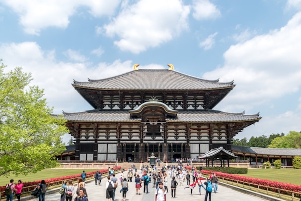 A large, traditional Japanese temple with ornate architectural detailing stands prominently. The temple features a grand entrance with a multi-tiered roof. In front of the temple, a diverse group of people are walking and taking photos. The surrounding area is landscaped with green trees and red flowers, under a partly cloudy blue sky.