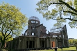 A partially destroyed building with a dome structure on top, surrounded by lush green trees and a clear blue sky. Scaffolding is visible inside the building, suggesting ongoing maintenance or preservation work.