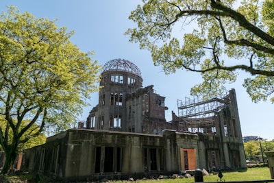 A partially destroyed building with a dome structure on top, surrounded by lush green trees and a clear blue sky. Scaffolding is visible inside the building, suggesting ongoing maintenance or preservation work.