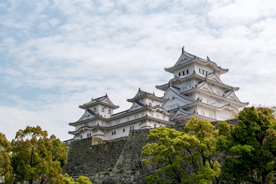 white and gray concrete castle surrounded by green trees under white clouds during daytime in Himeji Castle Japan