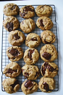 A batch of freshly baked chocolate chip cookies is cooling on a black wire rack. The cookies are golden brown with large, molten chocolate chunks visible on top, giving them a homemade and inviting appearance.