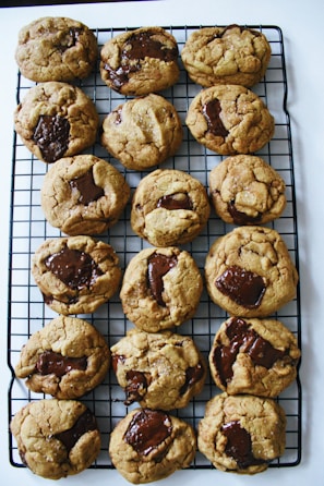 A batch of freshly baked chocolate chip cookies is cooling on a black wire rack. The cookies are golden brown with large, molten chocolate chunks visible on top, giving them a homemade and inviting appearance.