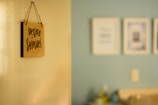 A blurred photograph of a light-colored wall with a hanging sign that reads 'Pedro Samuel' in dark, handwritten letters. The background shows a slightly out-of-focus interior room with a light blue wall and several framed pictures.