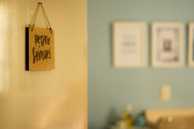 A blurred photograph of a light-colored wall with a hanging sign that reads 'Pedro Samuel' in dark, handwritten letters. The background shows a slightly out-of-focus interior room with a light blue wall and several framed pictures.