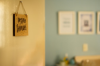 A blurred photograph of a light-colored wall with a hanging sign that reads 'Pedro Samuel' in dark, handwritten letters. The background shows a slightly out-of-focus interior room with a light blue wall and several framed pictures.