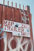 A rusty metal fence is topped with pointed spikes, and a white sign with red letters is attached to it. The sign reads 'RAP TRAILER REP' with additional numbers and symbols. There is graffiti in white and blue on the fence as well as on the sign. The image has a gritty, urban feel.