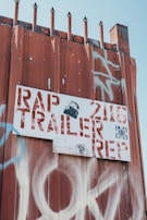 A rusty metal fence is topped with pointed spikes, and a white sign with red letters is attached to it. The sign reads 'RAP TRAILER REP' with additional numbers and symbols. There is graffiti in white and blue on the fence as well as on the sign. The image has a gritty, urban feel.