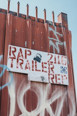 A rusty metal fence is topped with pointed spikes, and a white sign with red letters is attached to it. The sign reads 'RAP TRAILER REP' with additional numbers and symbols. There is graffiti in white and blue on the fence as well as on the sign. The image has a gritty, urban feel.