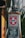 A metal pole with a label displaying a red cross and the word 'BONES,' indicating the brand 'Original Swiss.' Below it is a poster advertising an album titled 'The Obituary,' with music streaming service logos at the bottom. The pole is rusted and situated outdoors with leaves in the background.