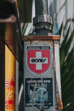 A metal pole with a label displaying a red cross and the word 'BONES,' indicating the brand 'Original Swiss.' Below it is a poster advertising an album titled 'The Obituary,' with music streaming service logos at the bottom. The pole is rusted and situated outdoors with leaves in the background.