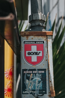 A metal pole with a label displaying a red cross and the word 'BONES,' indicating the brand 'Original Swiss.' Below it is a poster advertising an album titled 'The Obituary,' with music streaming service logos at the bottom. The pole is rusted and situated outdoors with leaves in the background.
