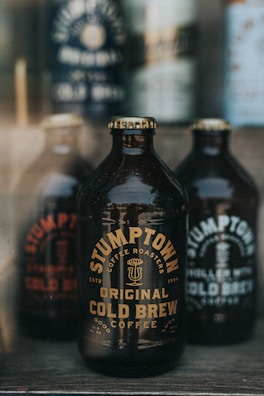 A dark, glass bottle labeled 'Stumptown Coffee Roasters Original Cold Brew Coffee' is prominently displayed in the foreground. Other similar bottles are blurred in the background, giving focus to the central bottle. The overall aesthetic is rustic and artisanal.