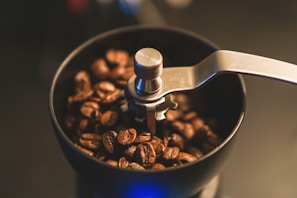 A manual coffee grinder filled with coffee beans, featuring a metal handle and gears visible at the top. The lighting is soft, highlighting the glossy texture of the coffee beans.