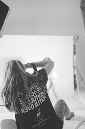 A black and white image of a person with long hair sitting on the floor and holding a camera, capturing a photograph in a studio setting. The person is wearing a T-shirt with text on the back and is positioned in front of a white backdrop. Lighting equipment is visible around the setting.