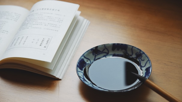 An open book with printed text in an Asian language lies on a wooden surface, accompanied by a porcelain dish containing black ink and a brush. The setting evokes a traditional and artistic ambiance.