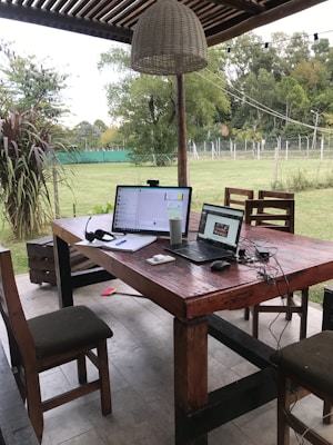 An outdoor workspace is set up with two laptops placed on a sturdy wooden table beneath a shaded area. Several chairs surround the table, and a large woven pendant light hangs above. The setting overlooks a grassy area and trees, creating a serene, natural environment. Various items such as a headset, a cup, and a notepad are scattered on the table.