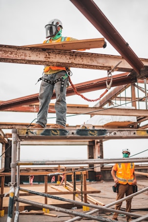 Two construction workers are on a building site. One worker stands on scaffolding, wearing safety gear including a helmet and harness, holding a wooden plank. The second worker is on the ground looking up, also in safety gear with a helmet and vest. The surroundings include metal beams and construction materials.
