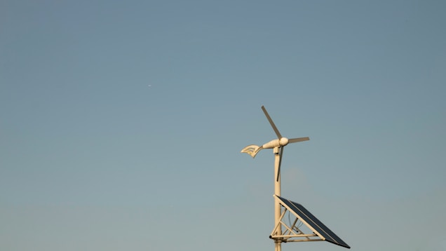 A wind turbine with a solar panel against a clear blue sky, placed on a simple metallic structure. The scene conveys a sense of clean, renewable energy and tranquility.