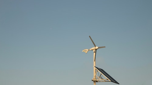 A wind turbine with a solar panel against a clear blue sky, placed on a simple metallic structure. The scene conveys a sense of clean, renewable energy and tranquility.