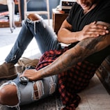 A person sitting on the floor with their legs stretched out, wearing ripped jeans, a black t-shirt, and a red and black checkered shirt tied around the waist. They have tattoos on their arm and are applying balm from a round container on their knee.