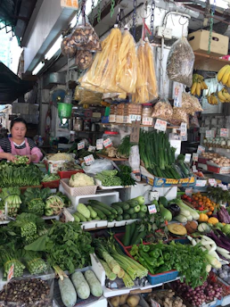 A bustling market stall displaying a variety of fresh vegetables and goods. The produce includes leafy greens, cucumbers, eggplants, gourds, peppers, and a variety of other vegetables. Some packaged goods and dried items hang from the ceiling. A woman stands to the left, arranging the produce or attending customers.