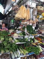 A bustling market stall displaying a variety of fresh vegetables and goods. The produce includes leafy greens, cucumbers, eggplants, gourds, peppers, and a variety of other vegetables. Some packaged goods and dried items hang from the ceiling. A woman stands to the left, arranging the produce or attending customers.