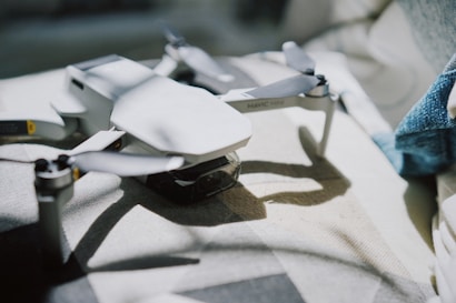 A sleek, white drone rests on a gray, textured surface, with soft lighting creating subtle shadows and highlights. The drone's propellers are prominently displayed, and part of a blue fabric is visible in the background.