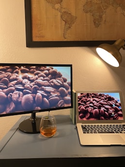 A desk setup featuring a computer monitor and a laptop side by side, both displaying close-up images of coffee beans. Above the desk, a framed vintage-style world map is mounted on the wall. A warm desk lamp is casting light on the scene, and there is a glass filled with a brown beverage placed on the desk.