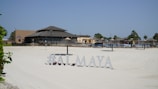 A sunlit beach area with silky white sand and a resort-style restaurant in the background. The sandy expanse leads to various thatched umbrellas and lounge chairs arranged for sunbathing. Palm trees are visible around the area, and a sign displays the text '#ALMAYA' near the forefront.