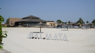 A sunlit beach area with silky white sand and a resort-style restaurant in the background. The sandy expanse leads to various thatched umbrellas and lounge chairs arranged for sunbathing. Palm trees are visible around the area, and a sign displays the text '#ALMAYA' near the forefront.